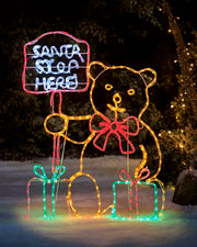 Pre-lit teddy bear Christmas silhouette holding a “Santa Stop Here” sign, with a large red bow and two illuminated presents at its feet, displayed outdoors in a snowy setting
