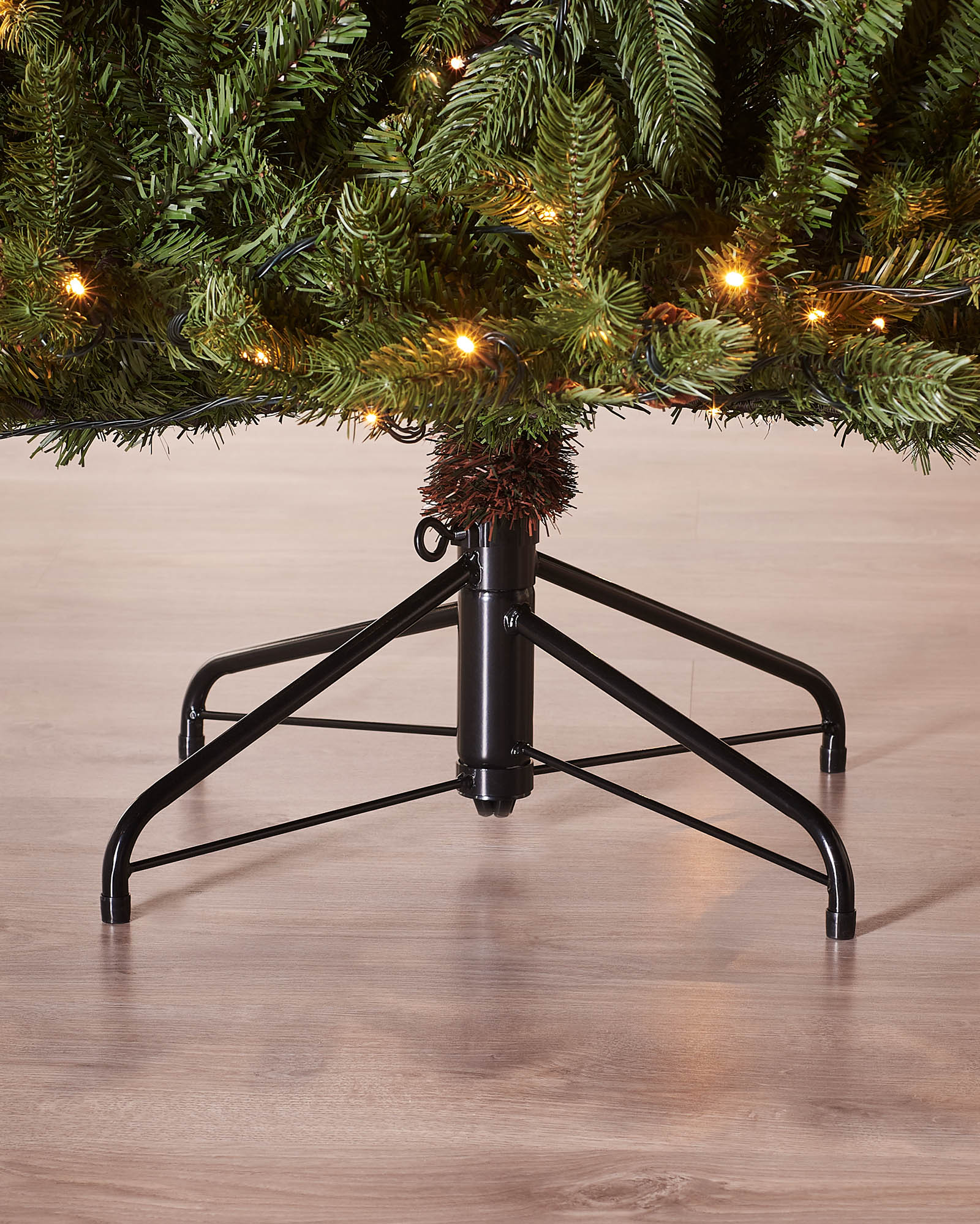 Close-up of the base of the Craford Pine Christmas tree with warm white lights, lower branches and a black metal tree stand on a wooden floor.