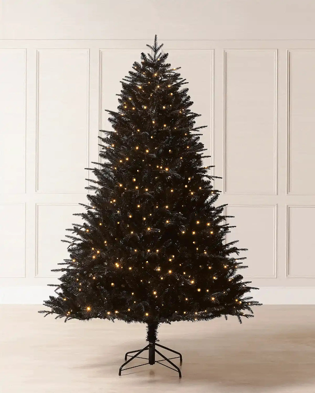 Full image of 7ft pre-lit black pine tree with warm white lighting in front of white panelling on light wood flooring