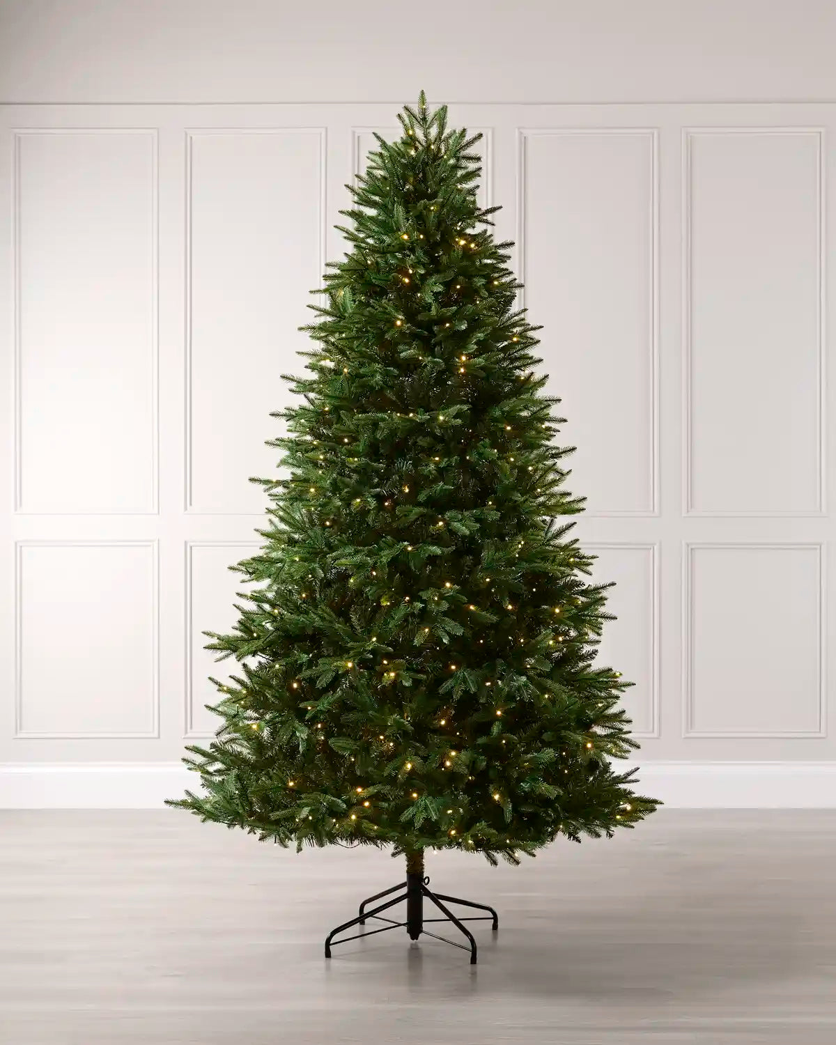 A tall, realistic artificial Christmas tree with warm white lights, standing on a black metal stand against a white paneled wall and light wood floor.