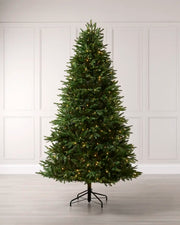 A tall, realistic artificial Christmas tree with warm white lights, standing on a black metal stand against a white paneled wall and light wood floor.