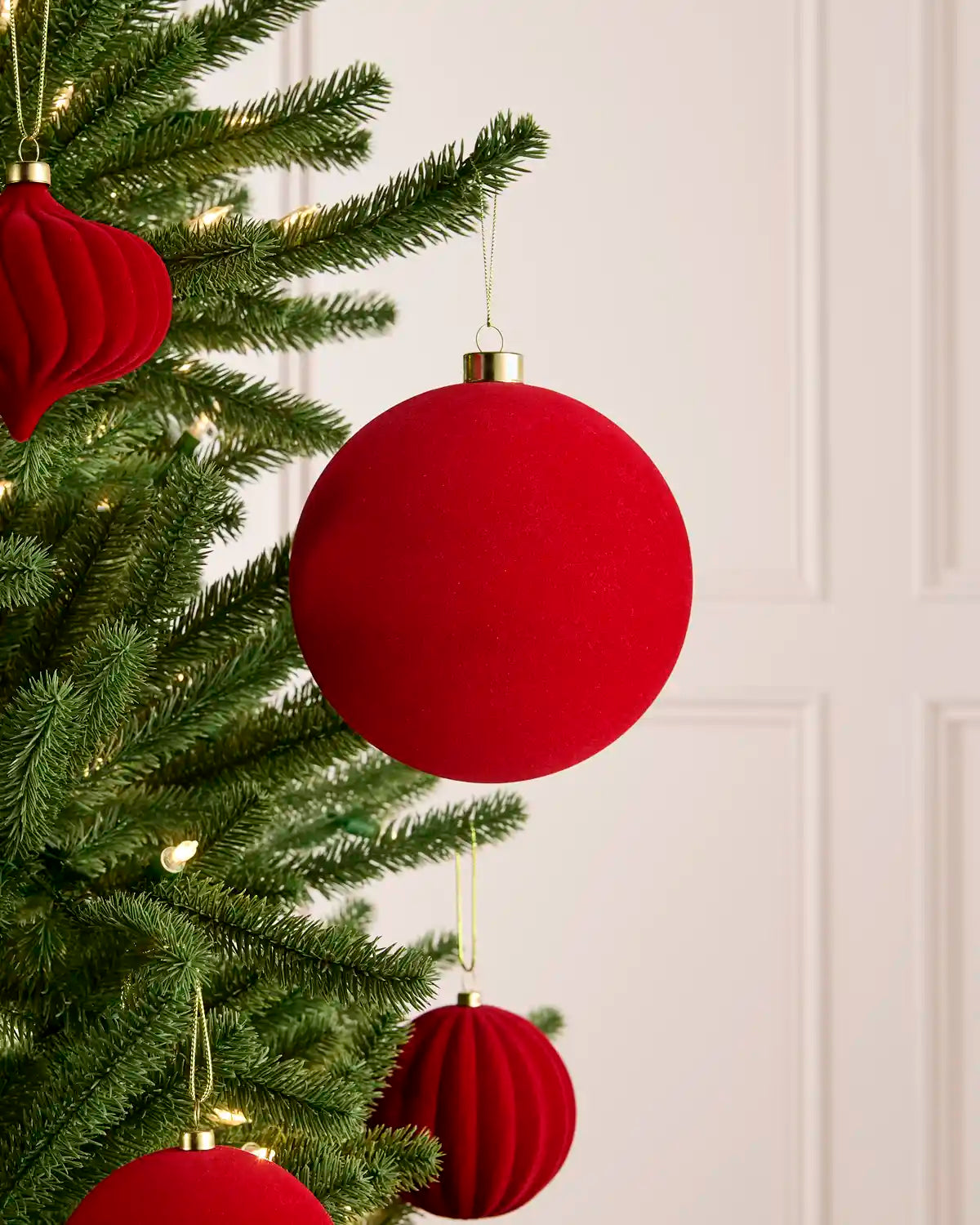 Red Christmas tree ornaments hanging on a green pine tree, including a large round ball ornament and pleated paper cone ornaments, against a soft cream paneled wall background.