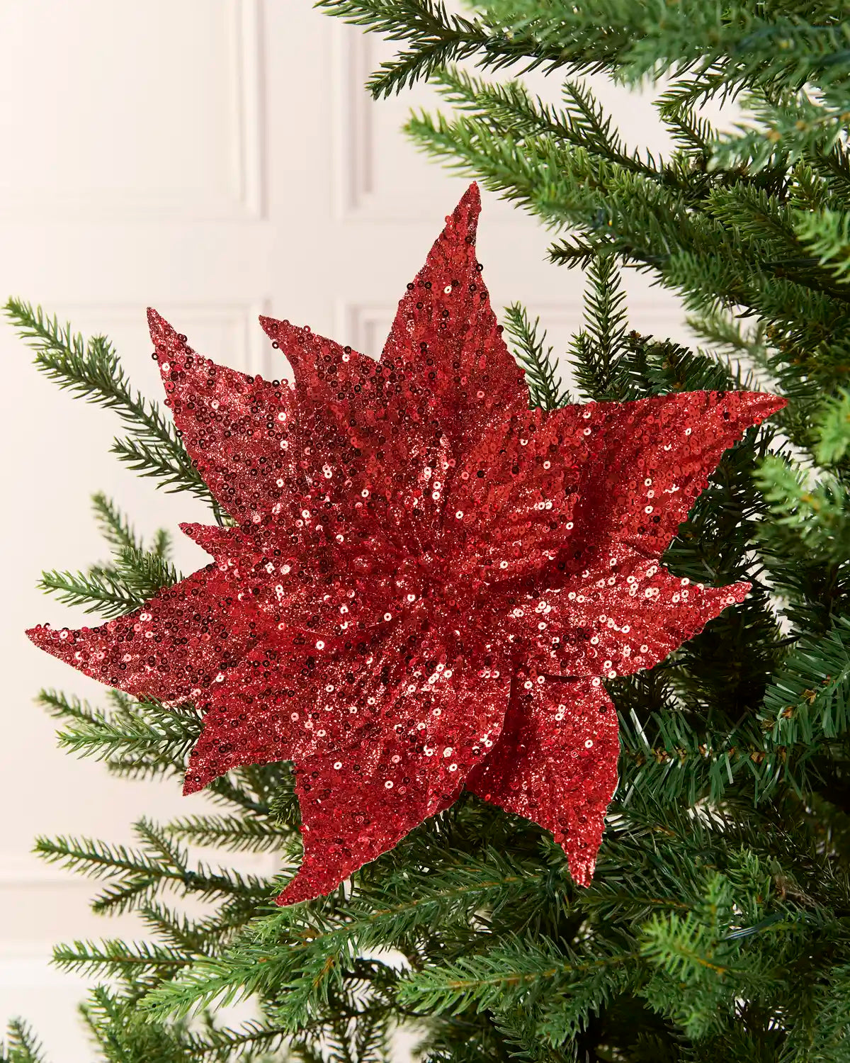 Red Sequin Artificial Poinsettia positioned in Christmas tree branches with a blurry cream background 