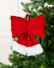 Red and white velvet Santa bow hanging from Christmas tree branch with blurry cream background.
