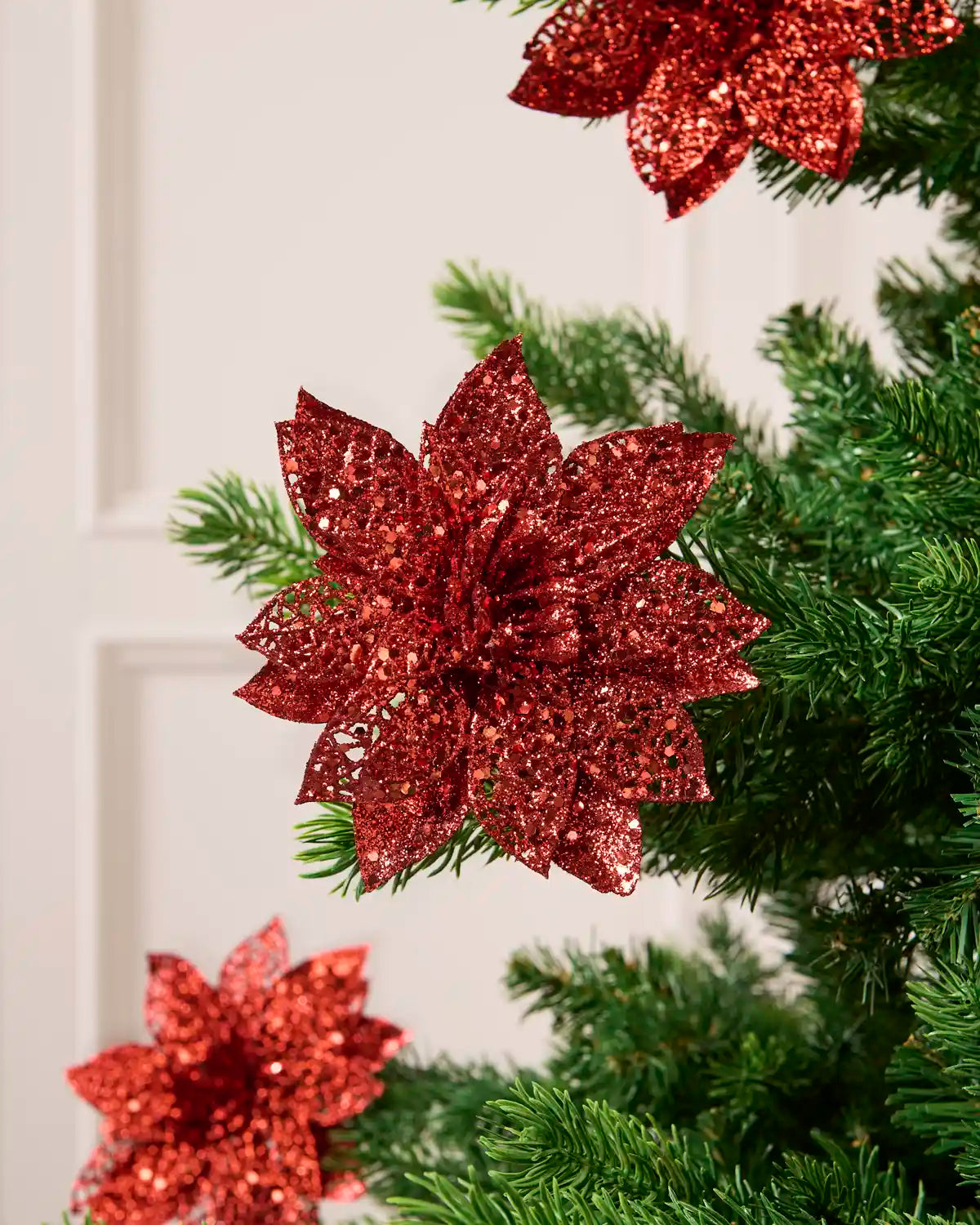 Red glitter artificial poinsettia flower with layered pointed petals, displayed among green Christmas tree.
