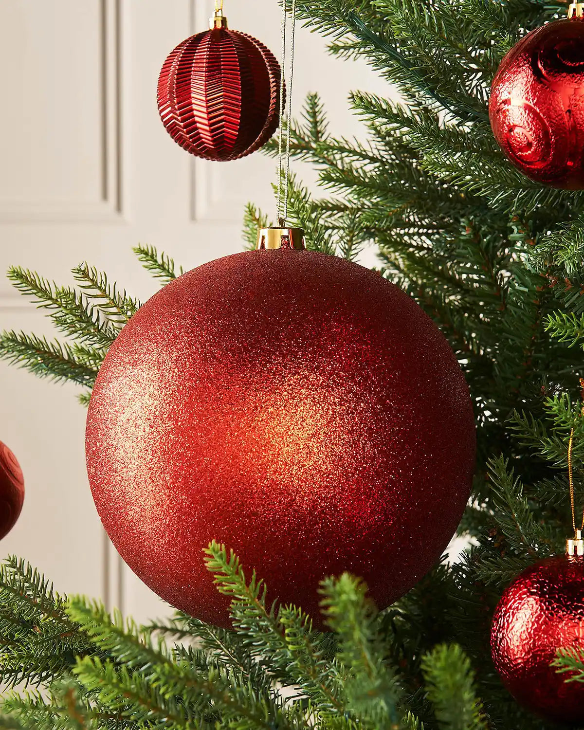 Red glittery oversized Christmas bauble ornament hanging on a tree branch with 4 smaller red baubles in background
