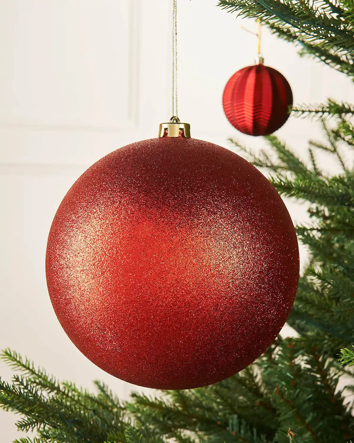 Red glittery oversized Christmas bauble ornament hanging on a tree branch with a smaller red bauble in background