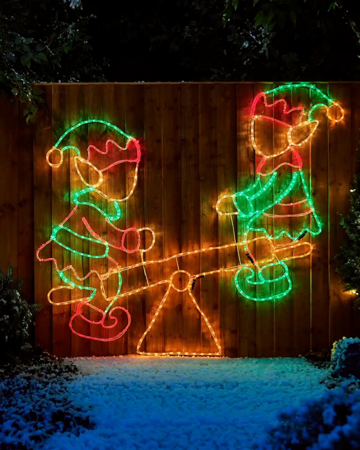 Red, green and warm white Elves on a seesaw Rope light silhouette  n a garden fence with snowy grass and trees in background