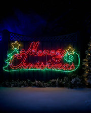 Red, green and yellow LED Merry Christmas Silhouette sign on a garden fence in a snowy garden  under a starry night sky