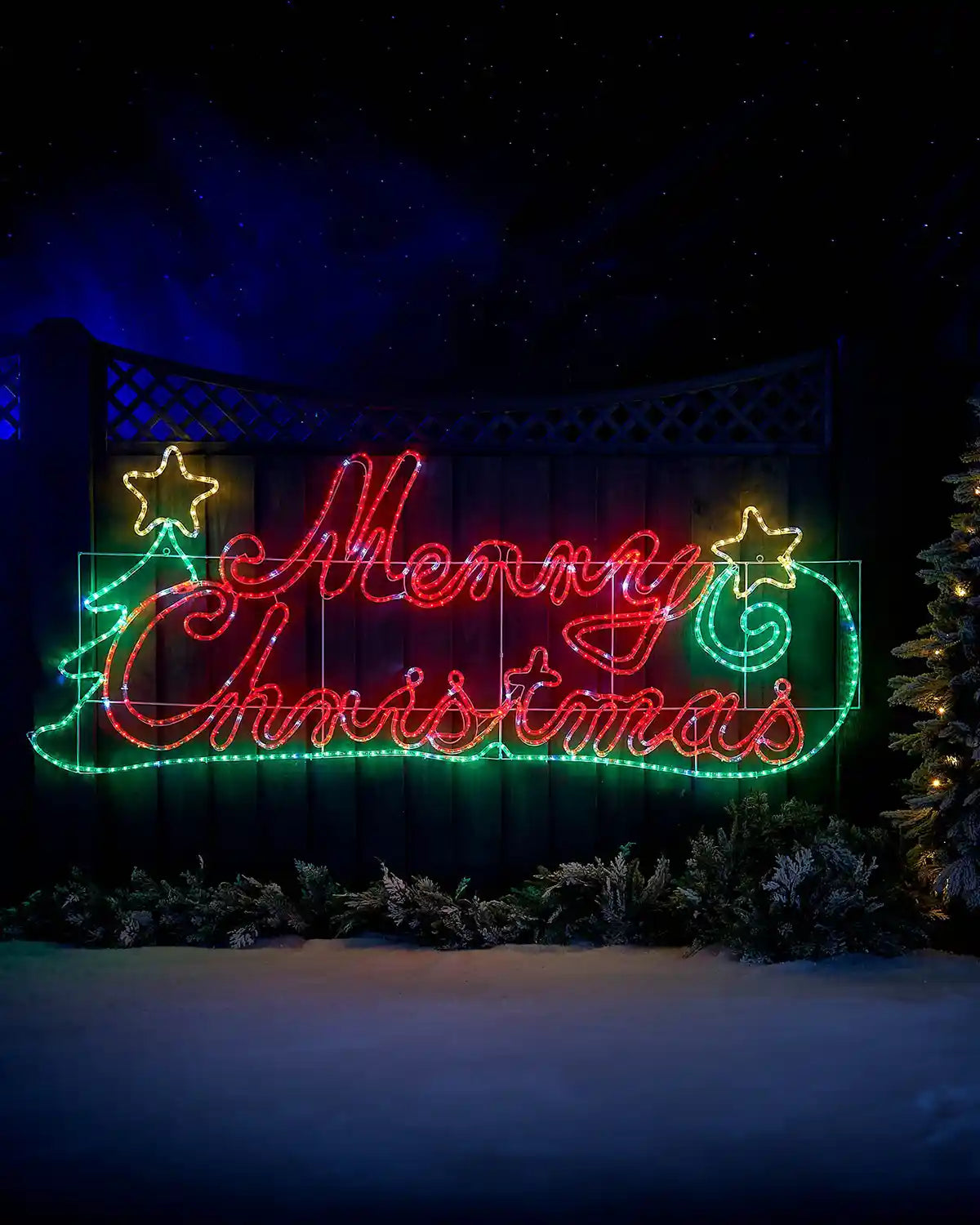 Red, green and yellow LED Merry Christmas Silhouette sign on a garden fence in a snowy garden  under a starry night sky