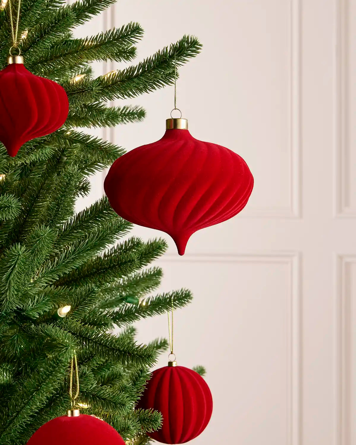 Red ribbed Christmas tree ornaments hanging on a green Christmas tree branch against a light cream paneled wall.
