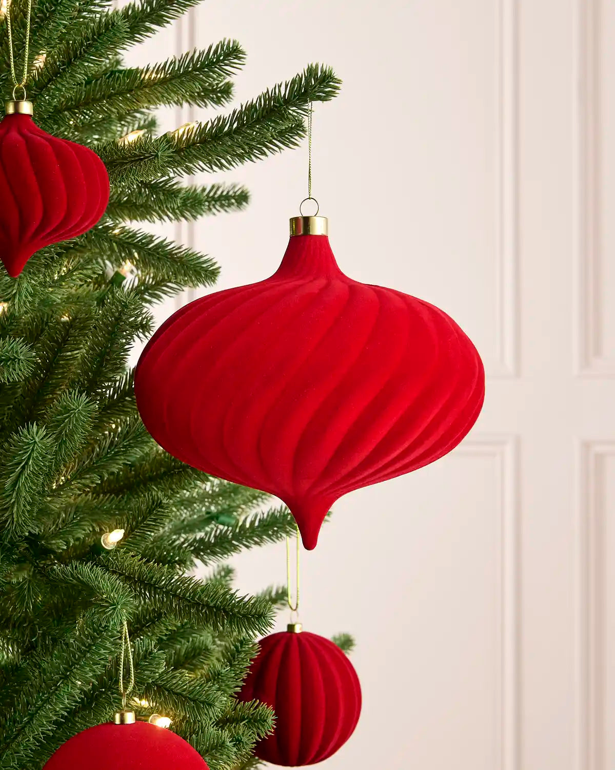 Red ribbed paper Christmas ornaments hanging on a green Christmas tree with warm white string lights against a light cream paneled wall.