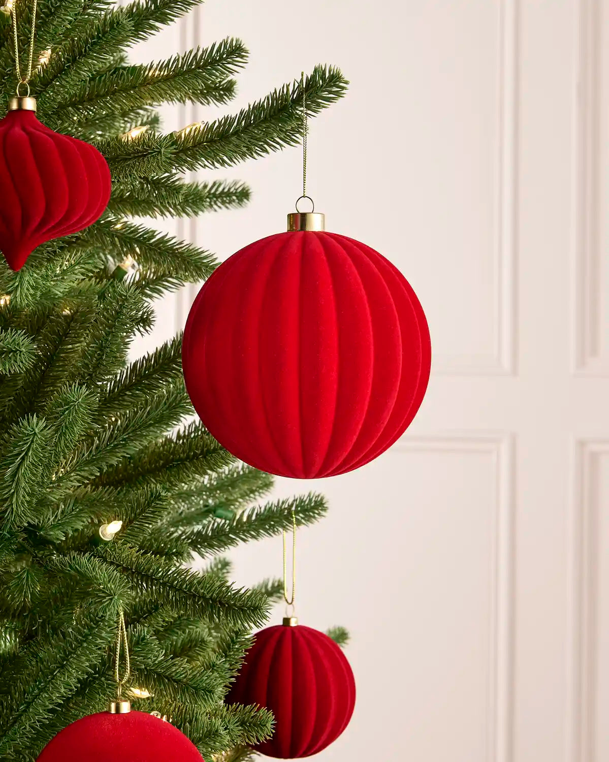 Red ribbed velvet Christmas tree ornaments hanging on a green pine tree branch against a white paneled wall.