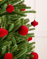 Red velvet Christmas tree ornaments hanging on a green artificial Christmas tree, with gold hanging loops against a light off-white panneled wall background.
