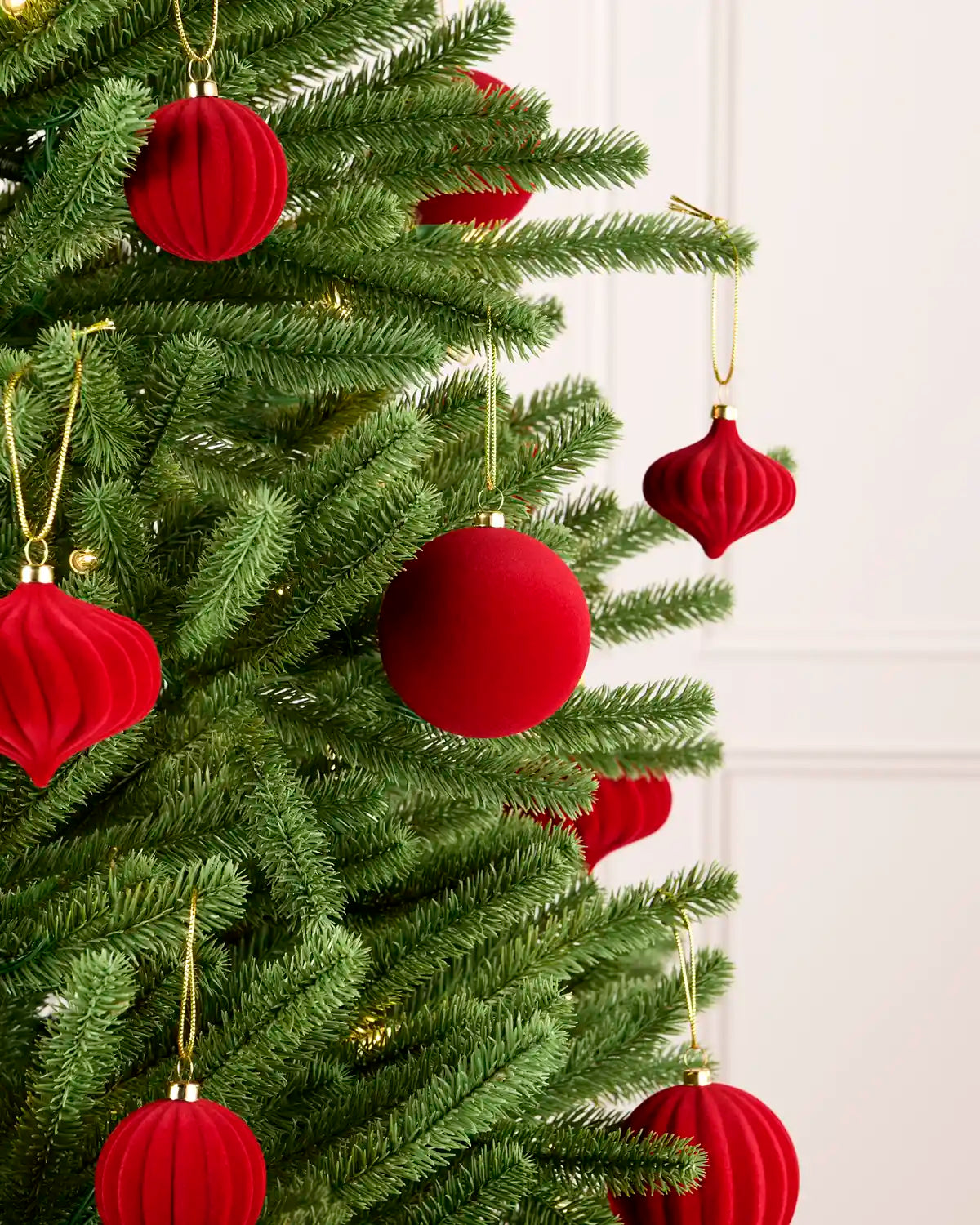 Red velvet Christmas tree ornaments hanging on a green artificial Christmas tree, with gold hanging loops against a light off-white panneled wall background.