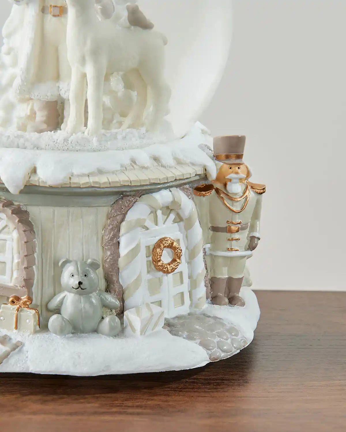 Right side close-up of musical Santa snow globe featuring silver teddy bear and cream/gold nutcracker character on  a dark wood surface in front of white wall