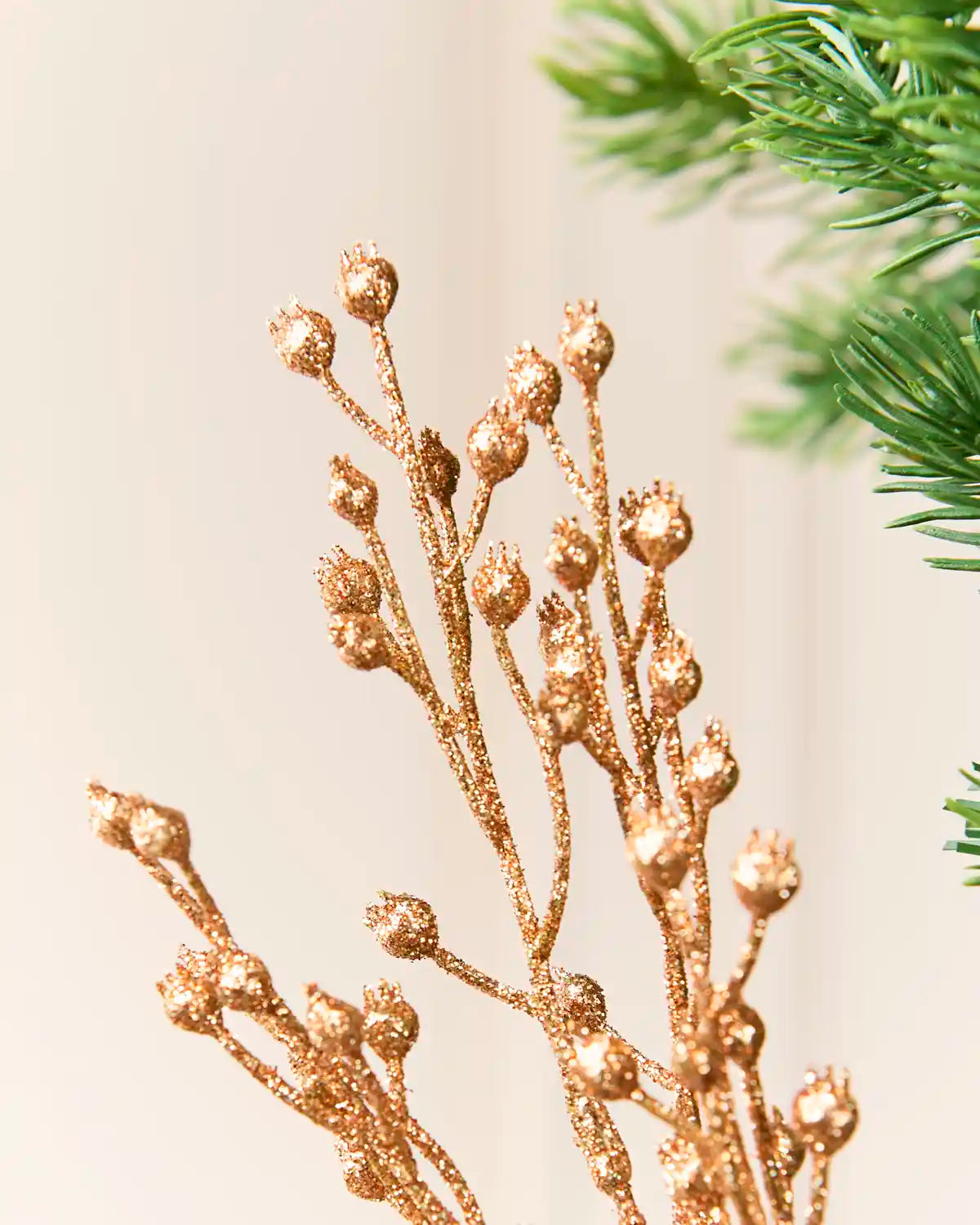 Rose Gold glitter artificial floral stems with textured buds, shown against a soft cream background with blurred green pine branches in the corner.