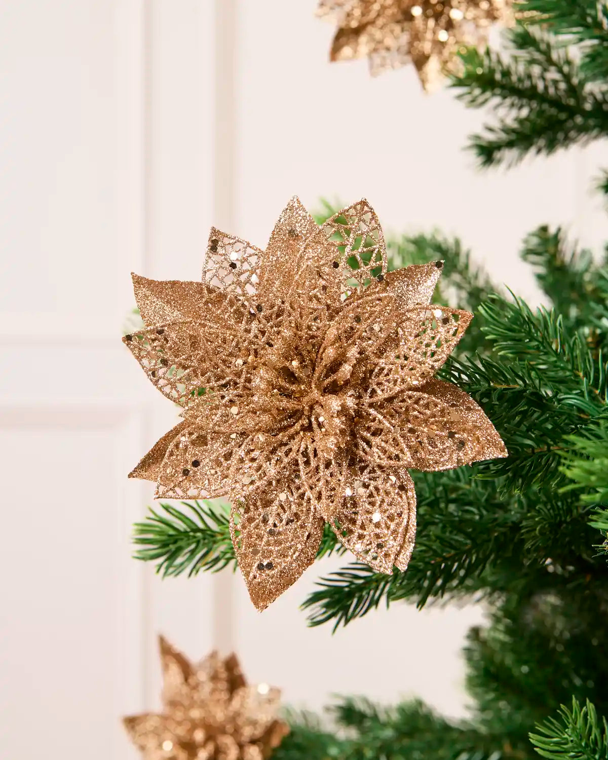 Rose Gold glitter artificial poinsettia flower with layered pointed petals, displayed among green Christmas tree.
