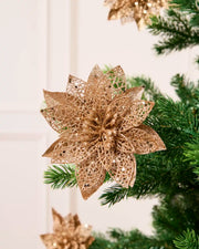Rose Gold glitter artificial poinsettia flower with layered pointed petals, displayed among green Christmas tree.
