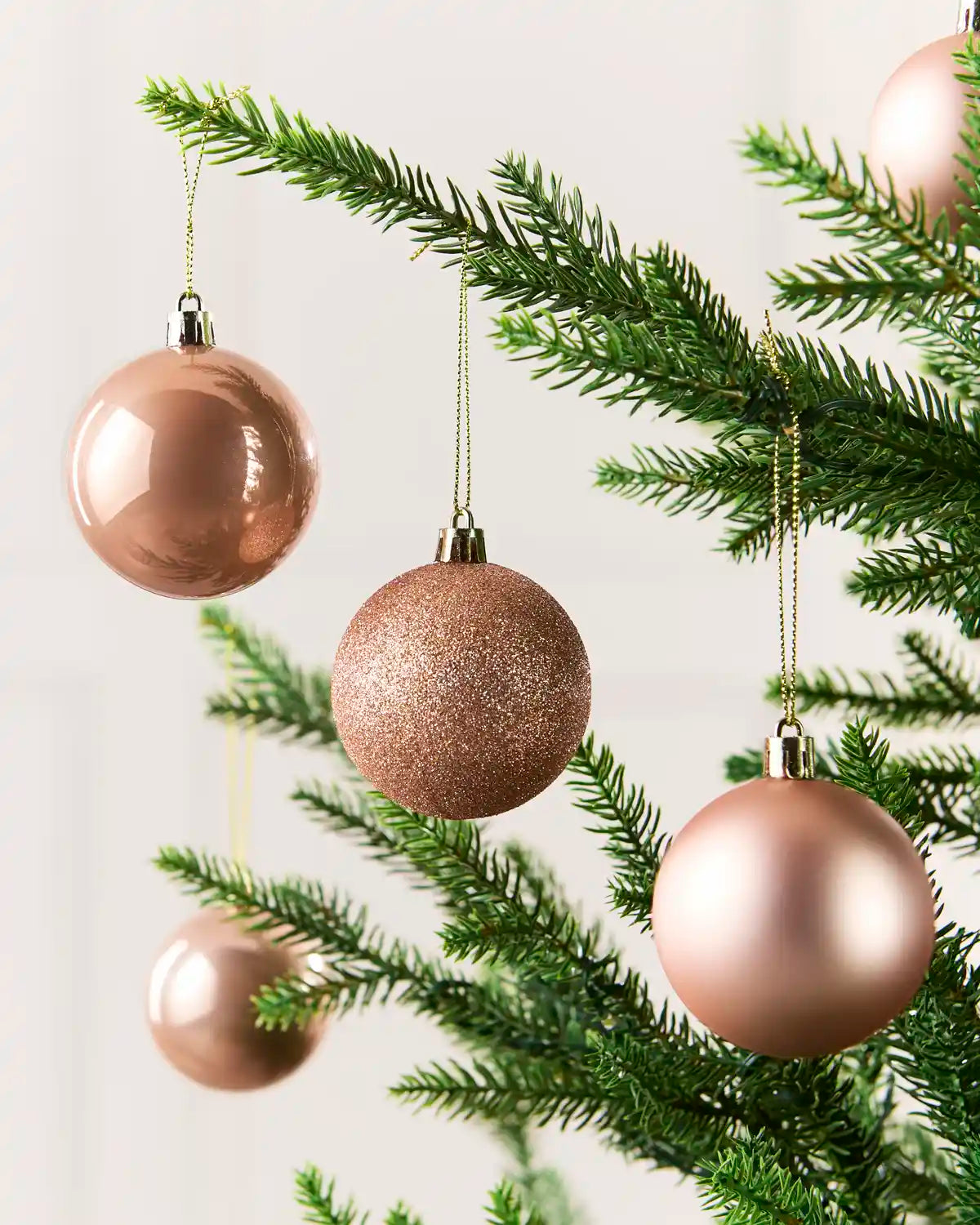 Rose gold Christmas baubles ornaments hanging on green pine branches against a light neutral background.
