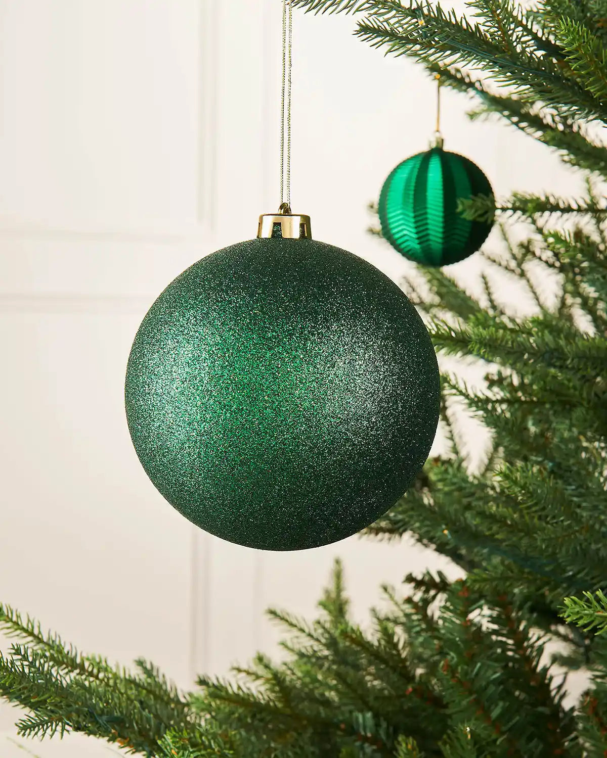 Sage glitter green oversized 15cm bauble on a Christmas tree branch with a smaller green bauble in the background