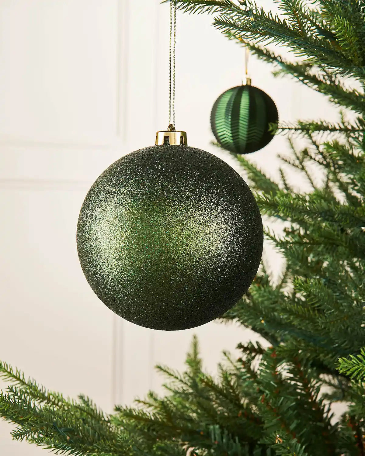 Sage green glitter oversized 15cm bauble on a Christmas tree branch with a smaller sage green bauble in the background