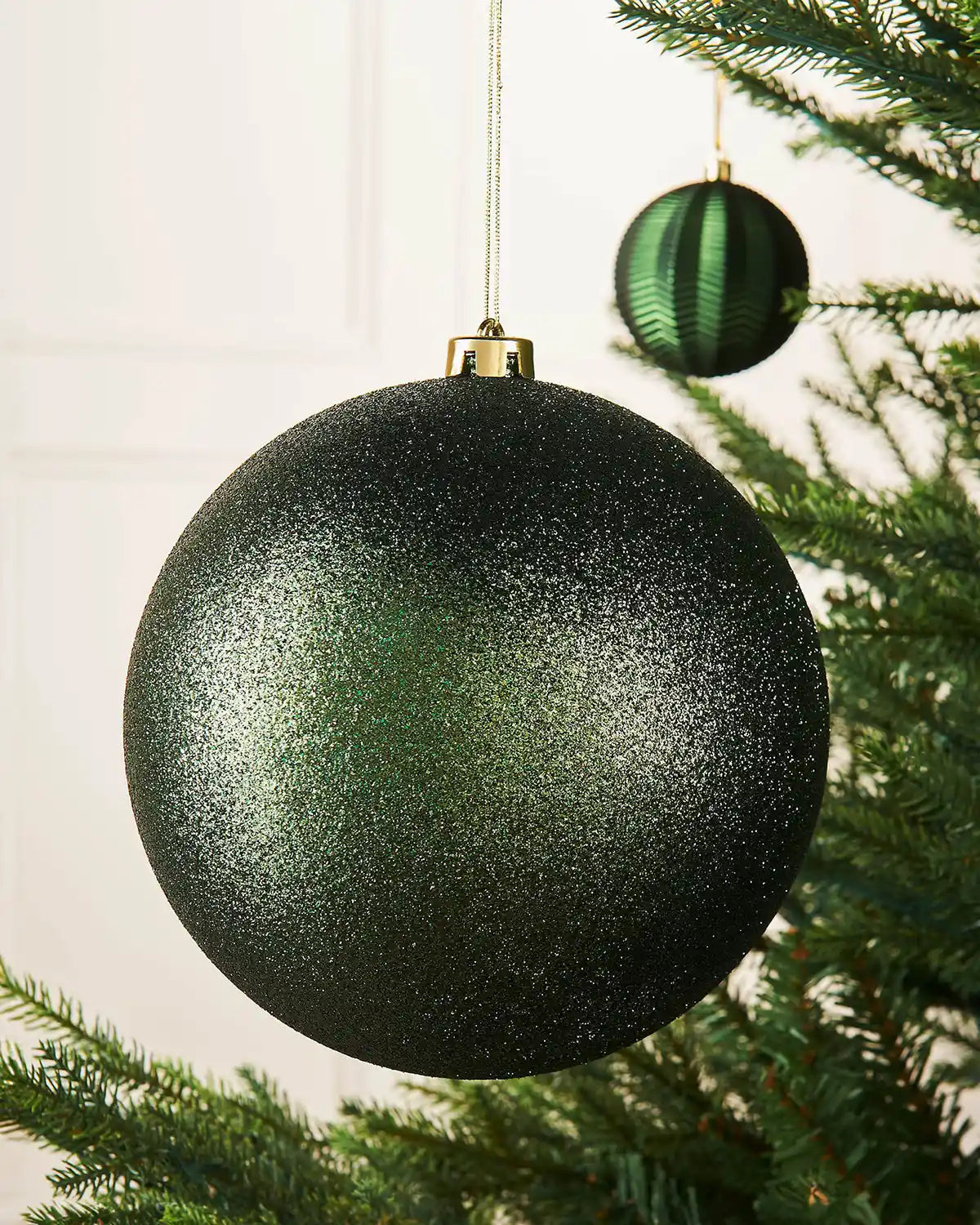 Sage green glitter oversized bauble on a Christmas tree branch with a smaller sage green bauble in the background