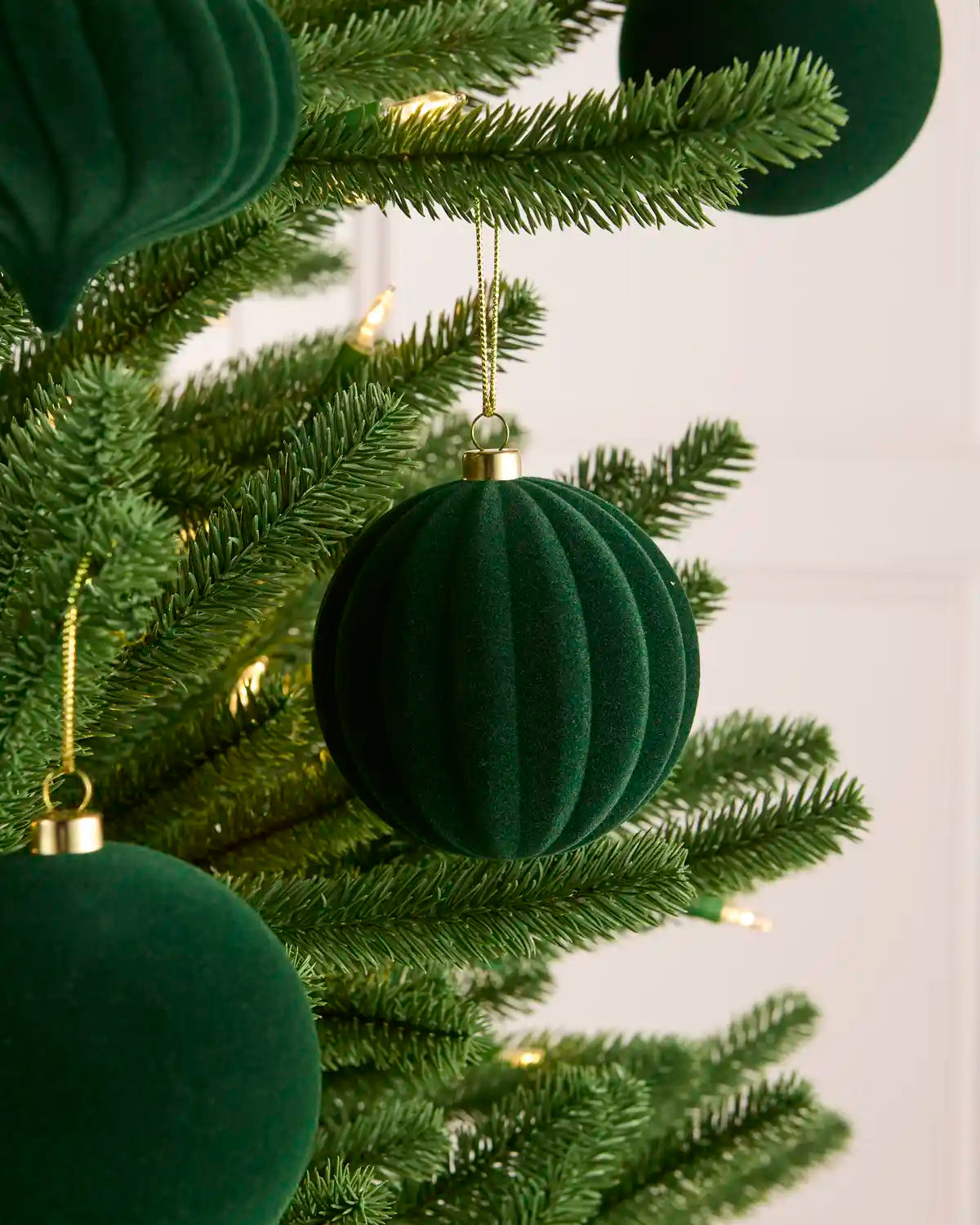 Sage green ribbed velvet Christmas tree ornaments hanging on a green artificial Christmas tree with warm white string lights against a light background.