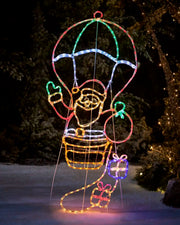 Santa in a hot air balloon rope light in red, blue, warm white and green silhouette with presents against a greenery background on snowy grass