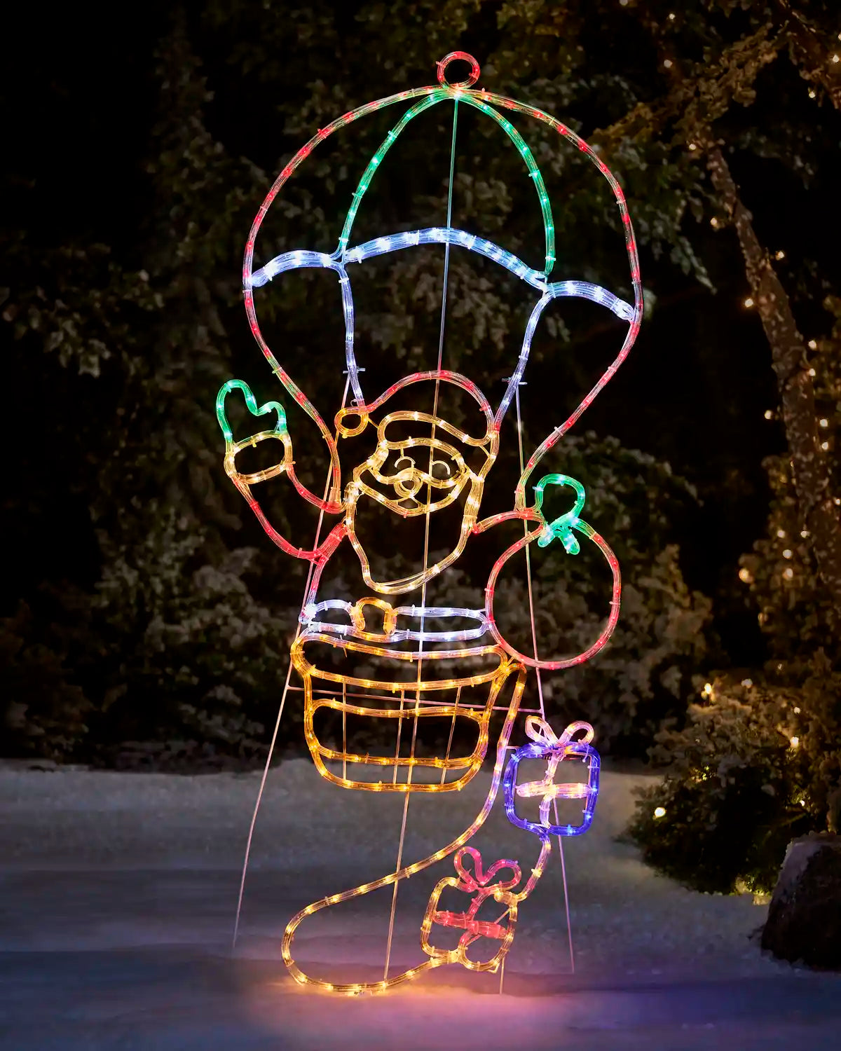 Santa in a hot air balloon rope light in red, blue, warm white and green silhouette with presents against a greenery background on snowy grass