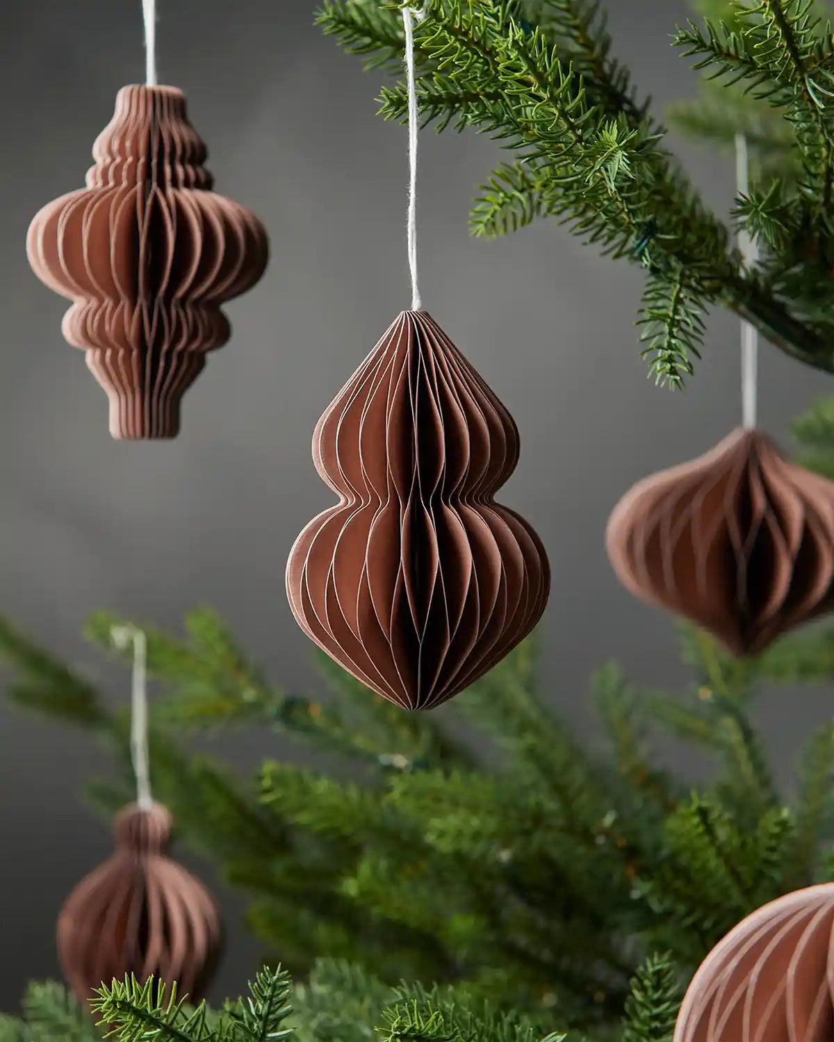 Scattered collection of 10cm mocha brown paper tree decorations hanging on Christmas tree in front of grey wall background