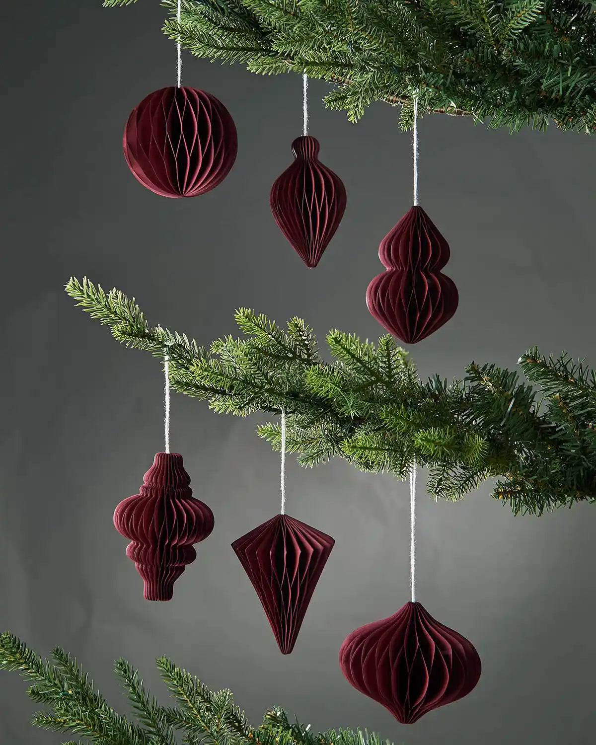 Set of 6 burgundy red paper Christmas tree decorations hanging on tree in front of grey background