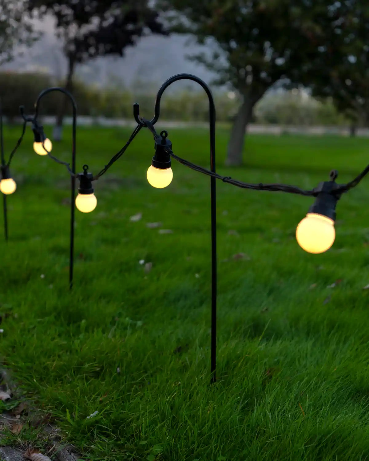 Shepherd’s hook garden stake with a curved black metal top supporting outdoor string lights with garden background
