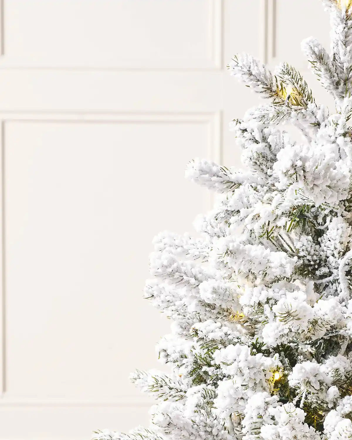 Side close up view of a Pre-Lit Snow Flocked Potted Christmas Tree, 3 ft against a cream panel wall