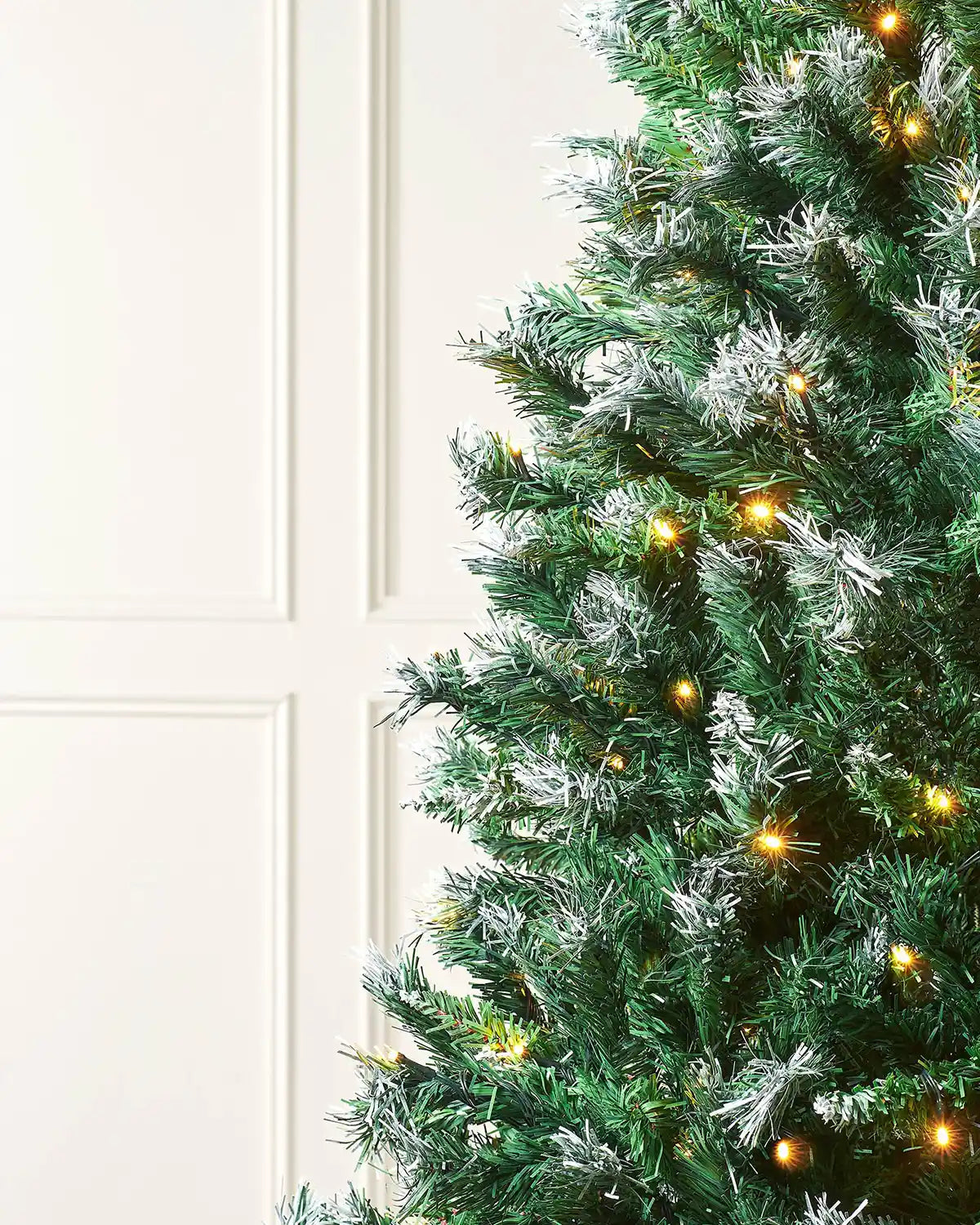 Side view of a Pre-Lit Frosted Mixed Pine Christmas Tree, 7ft with warm white lights against a cream panel wall
