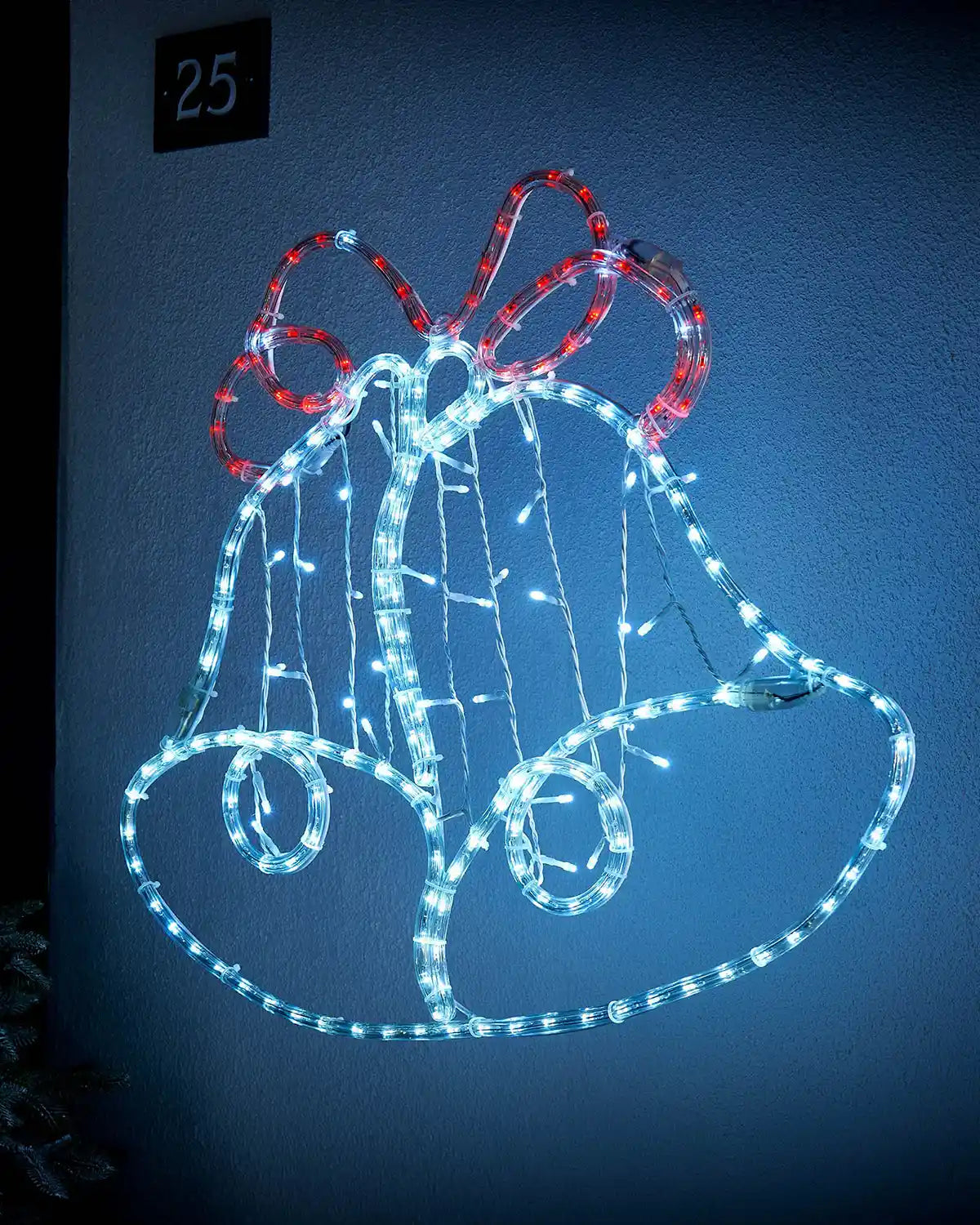 Side view of a White and Red Light up Christmas bell silhouette on a light coloured wall outside with a small Christmas tree in corner
