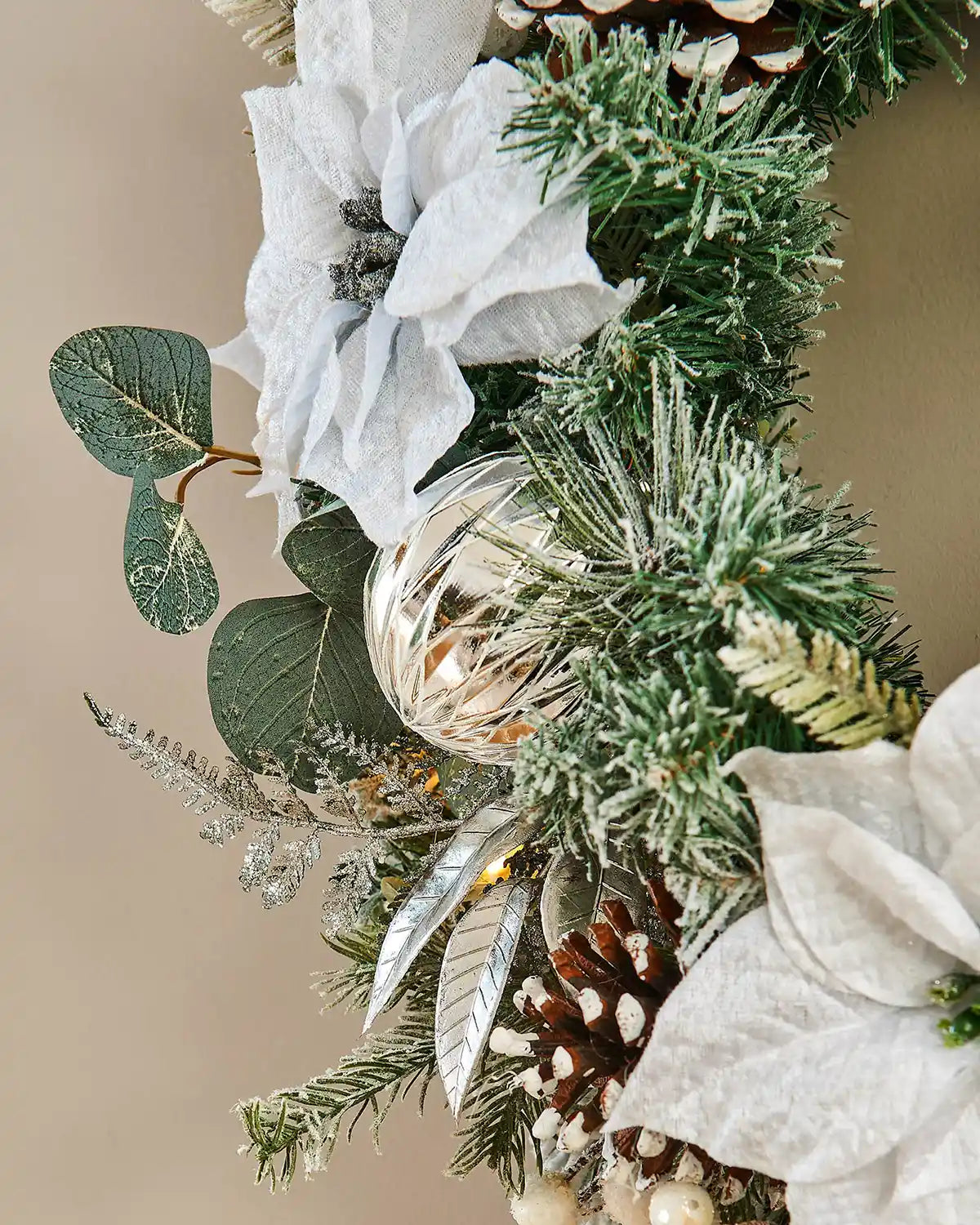 Side view of decorative wreath with greenery, white flowers, and pinecones on a beige background