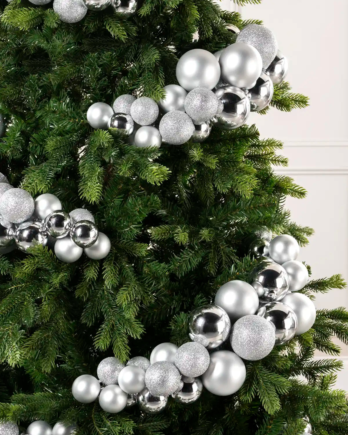 Silver and white Christmas tree bauble garland ornaments, including shiny metallic balls, matte silver spheres, and glittery ornaments, arranged on a green artificial Christmas tree.
