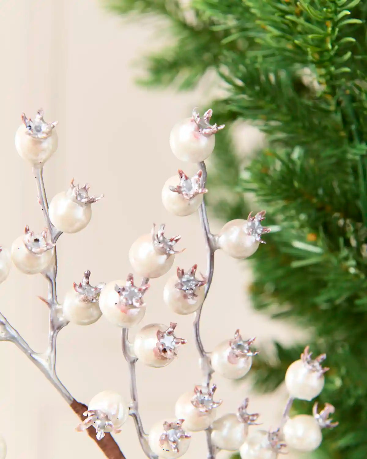 Silver decorative pick adorned with faux white pearl berries and small silver starburst accents, shown against a soft beige background with blurred green foliage on the right.