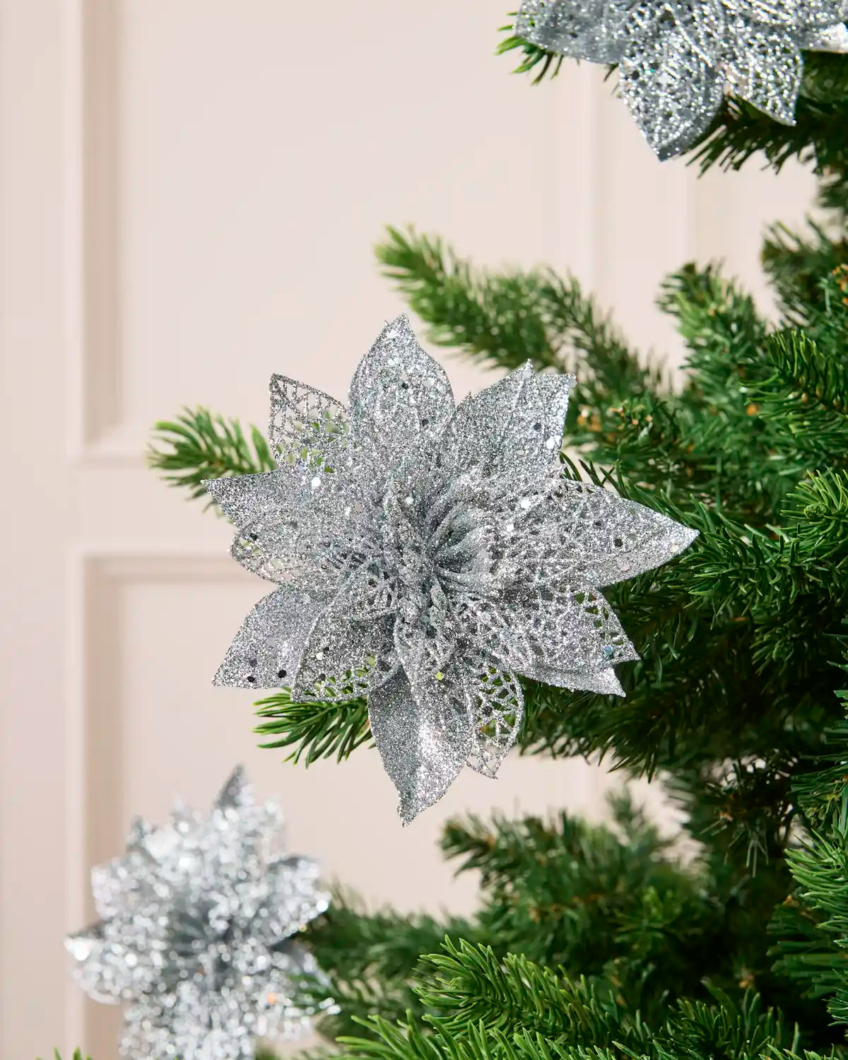 Silver glitter artificial poinsettia flower with layered pointed petals, displayed among green Christmas tree.
