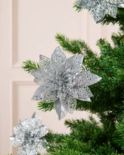 Silver glitter artificial poinsettia flower with layered pointed petals, displayed among green Christmas tree.
