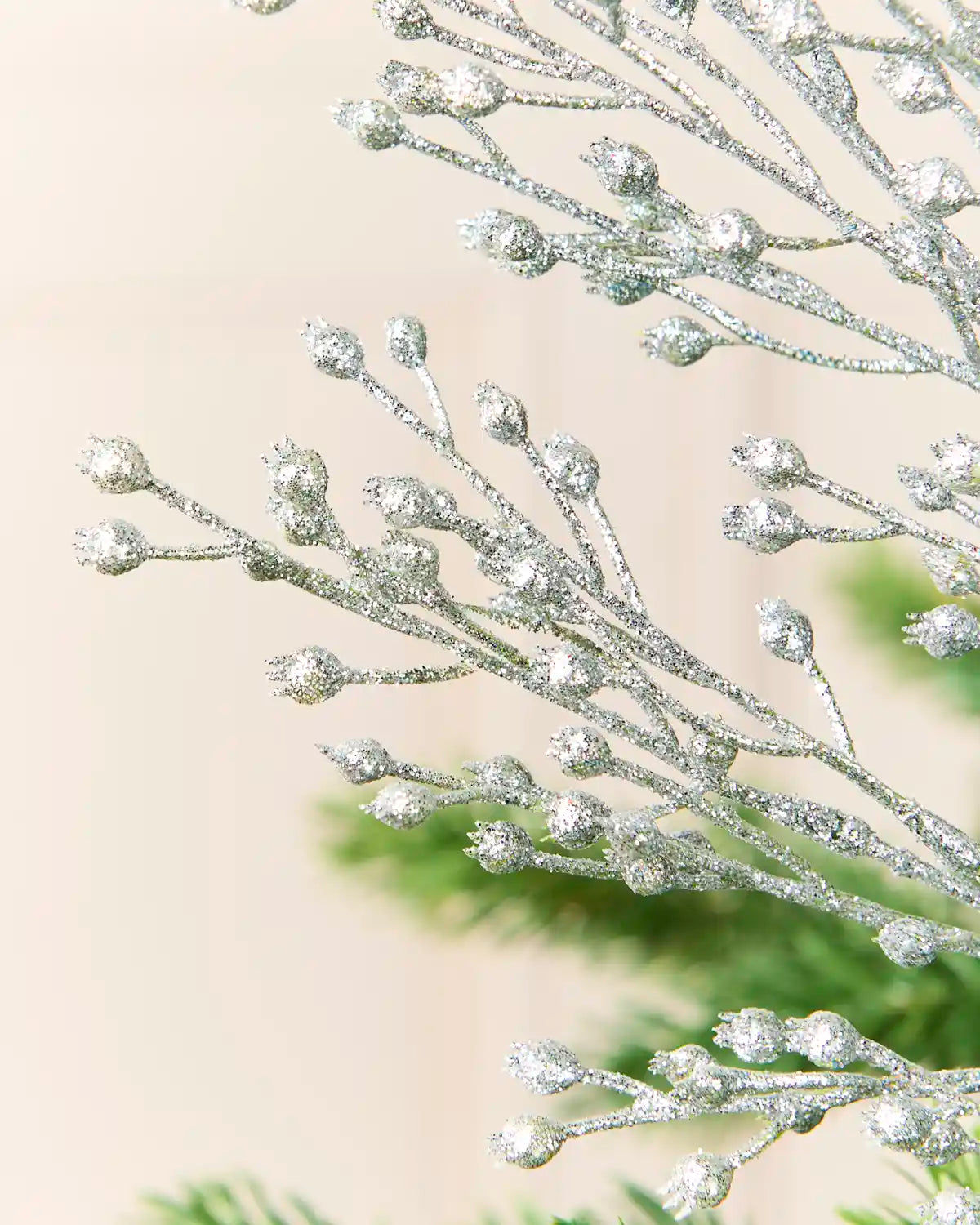 Silver glitter berry spray with small rounded buds, shown close up against a soft beige background with blurred green foliage at the bottom.
