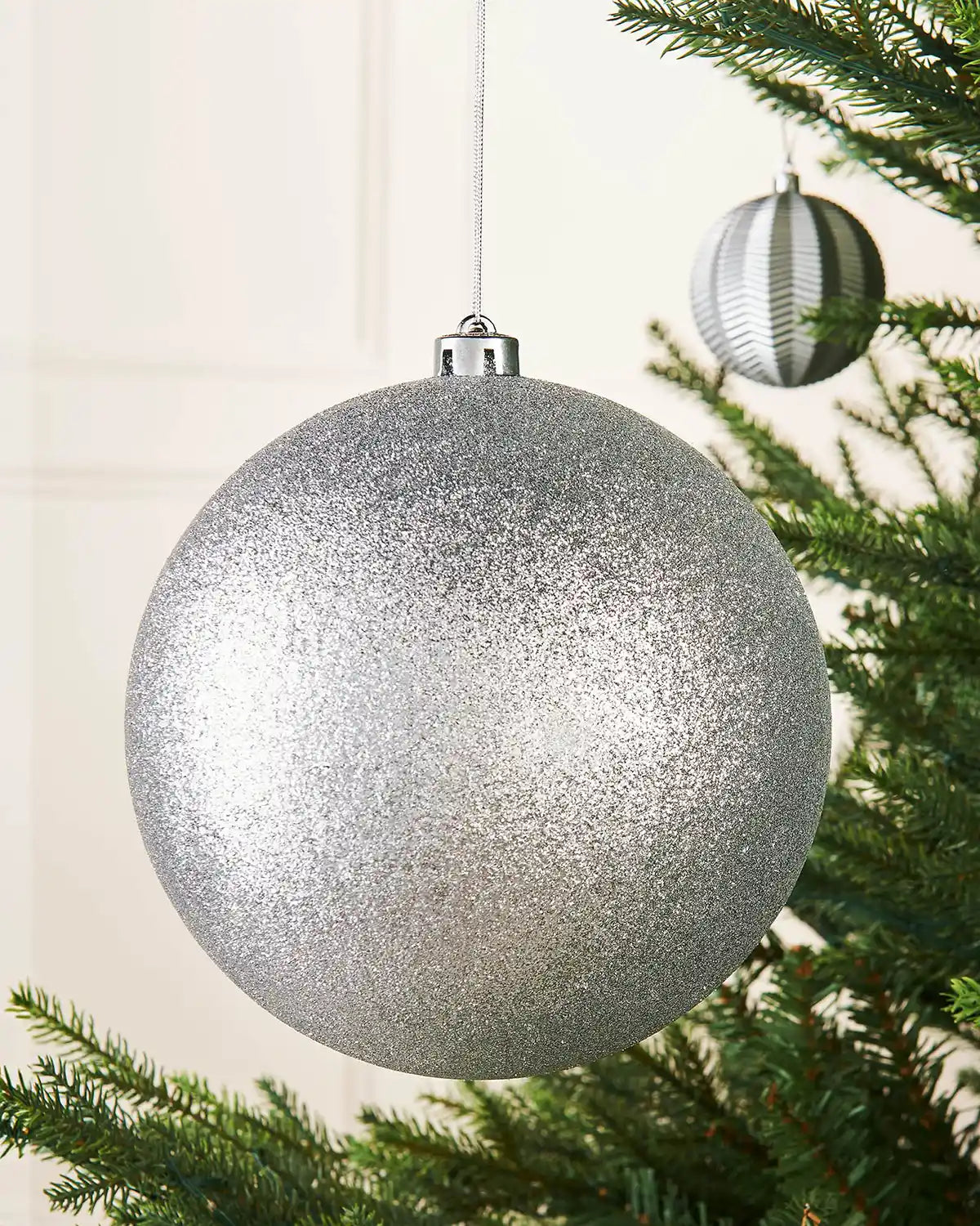 Silver glittery Christmas bauble hanging on a tree branch with a smaller silver bauble in background