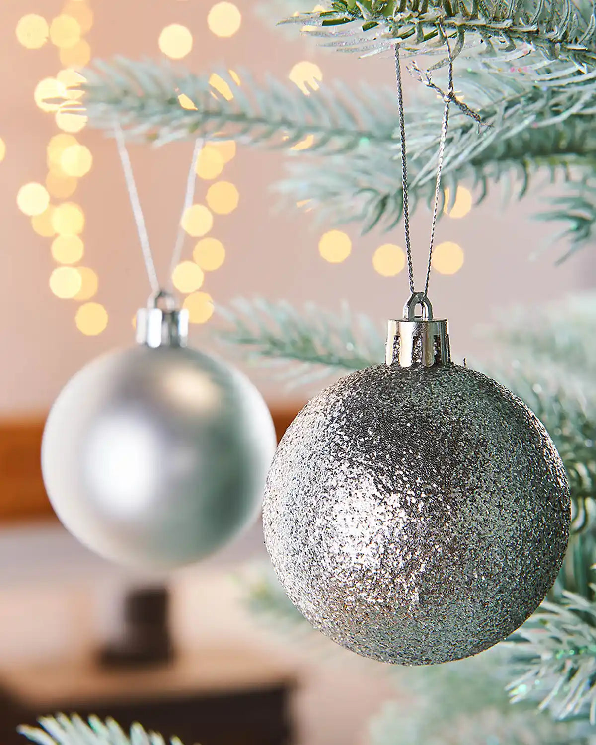 Silver glittery Christmas baubles hanging on a tree branch with blurred lights in the background