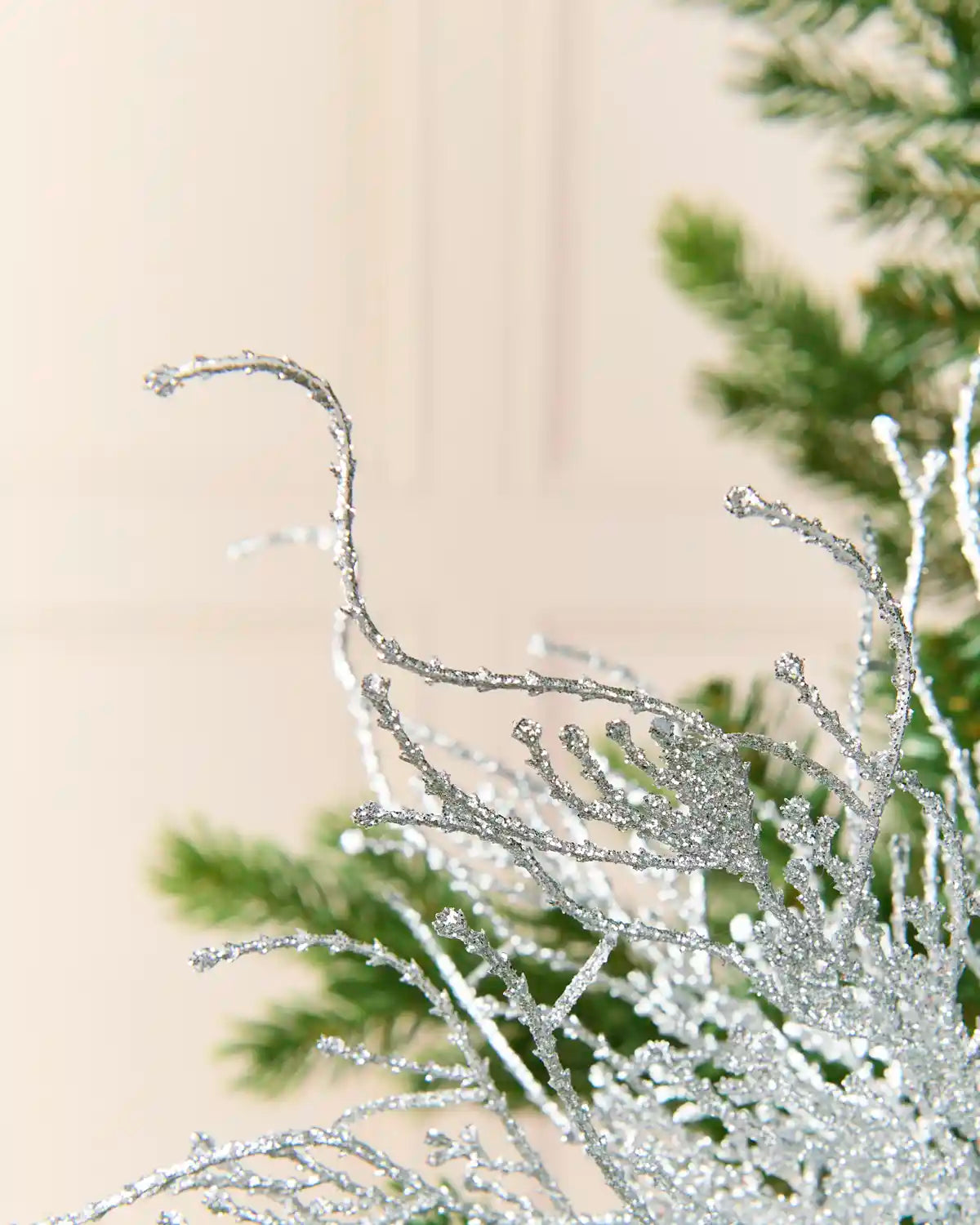 Silver glittery decorative branches with a frosted, icy texture, shown in close-up against a soft beige background with blurred green pine branches.