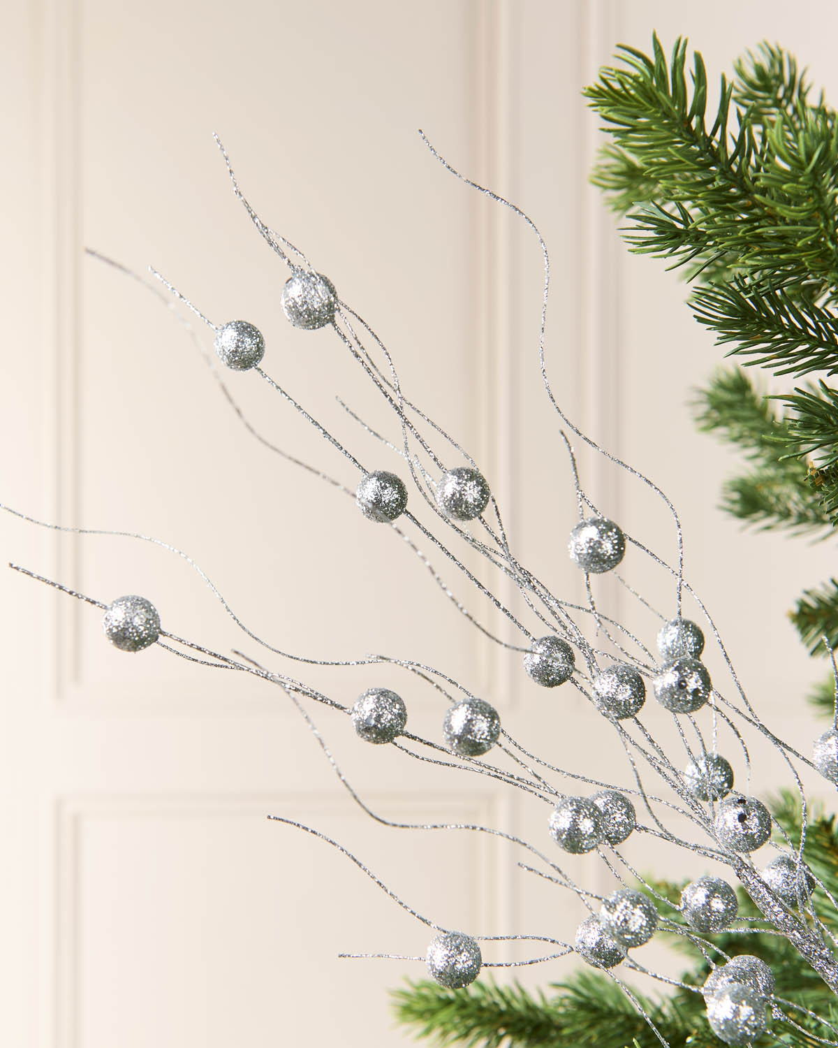 Silver glittery decorative branches with round metallic ornaments, shown draped over green pine tree branches against a soft cream paneled background.