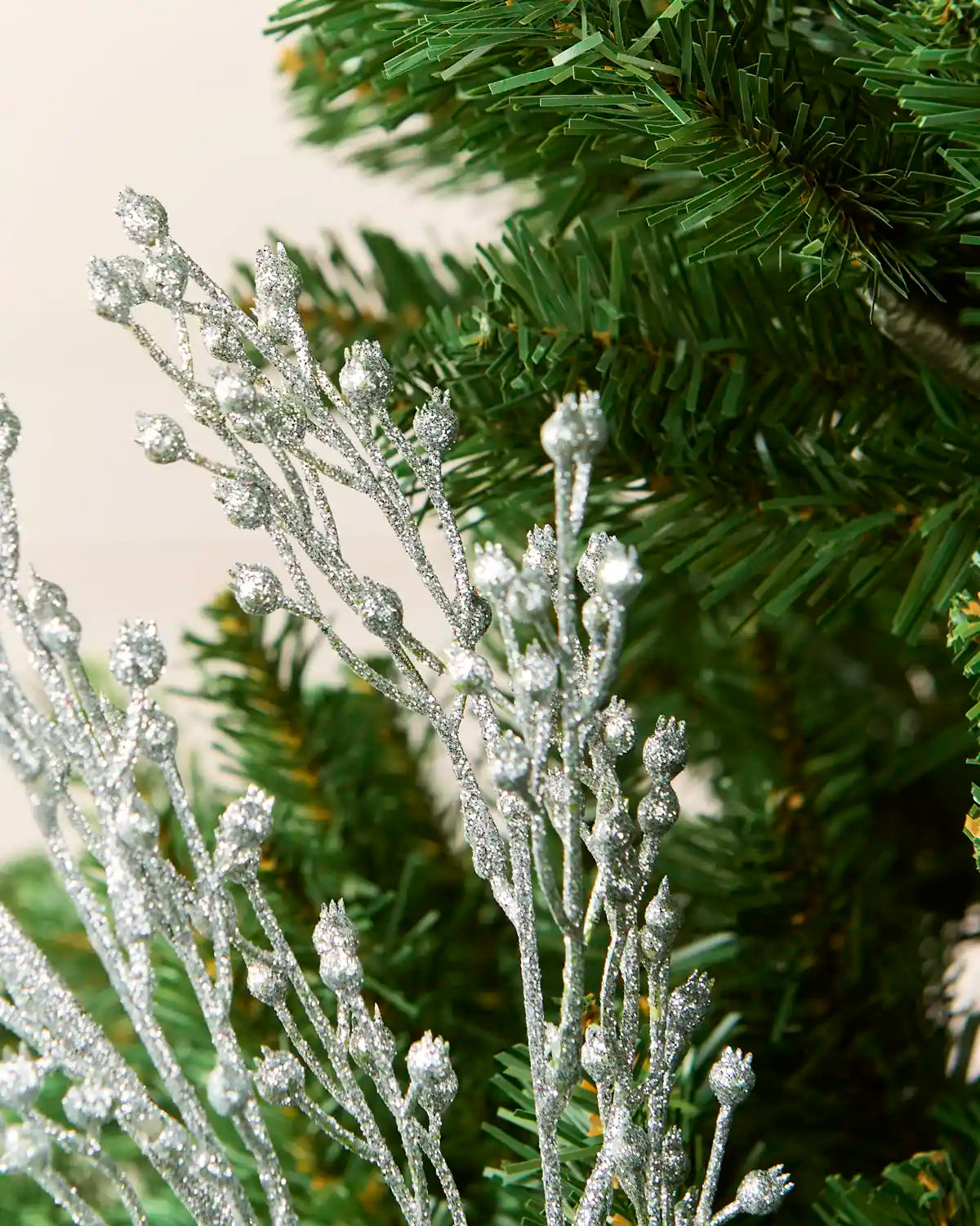 Silver glittery decorative spray ornament with frosted buds, shown close-up against green artificial pine needles and a light neutral background.