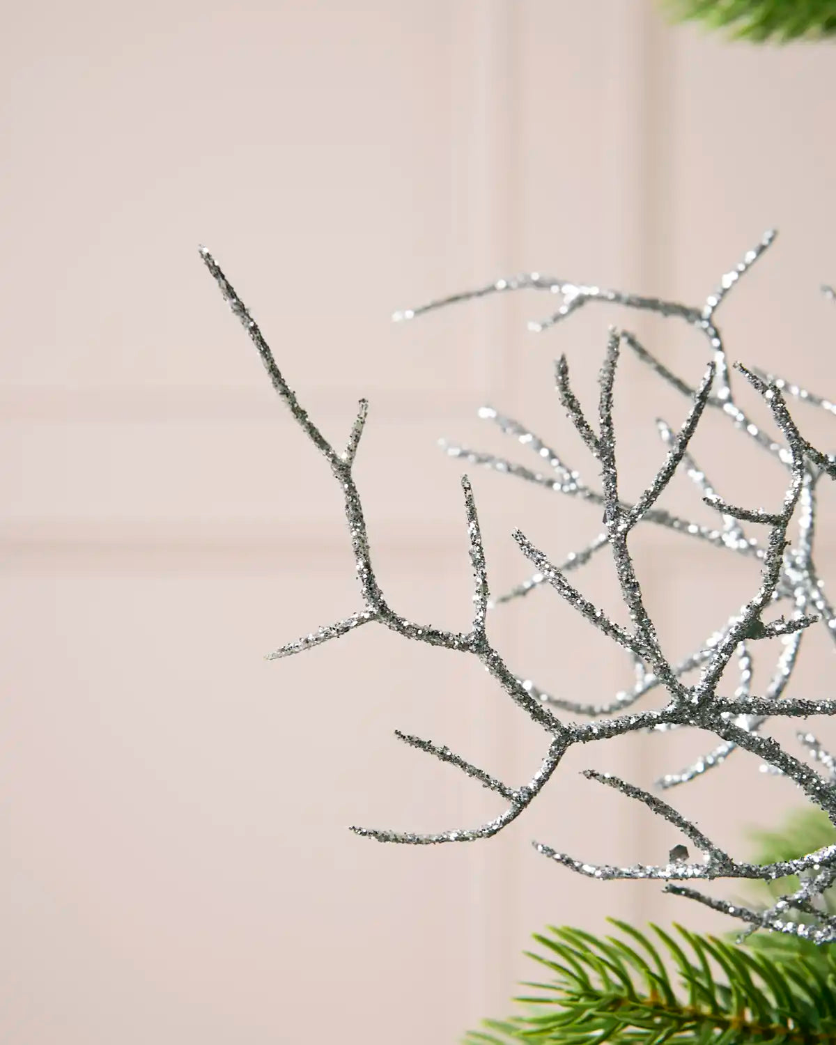 Silver glittery decorative branches with a sparkling finish, shown against a soft beige background with hints of green pine foliage at the bottom right.