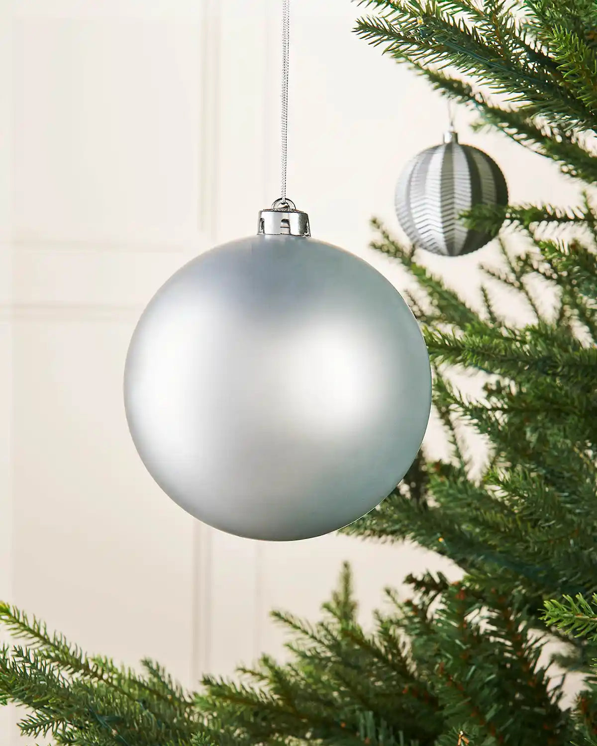 Silver matt oversized 15cm bauble on a Christmas tree branch with a smaller silver bauble in background