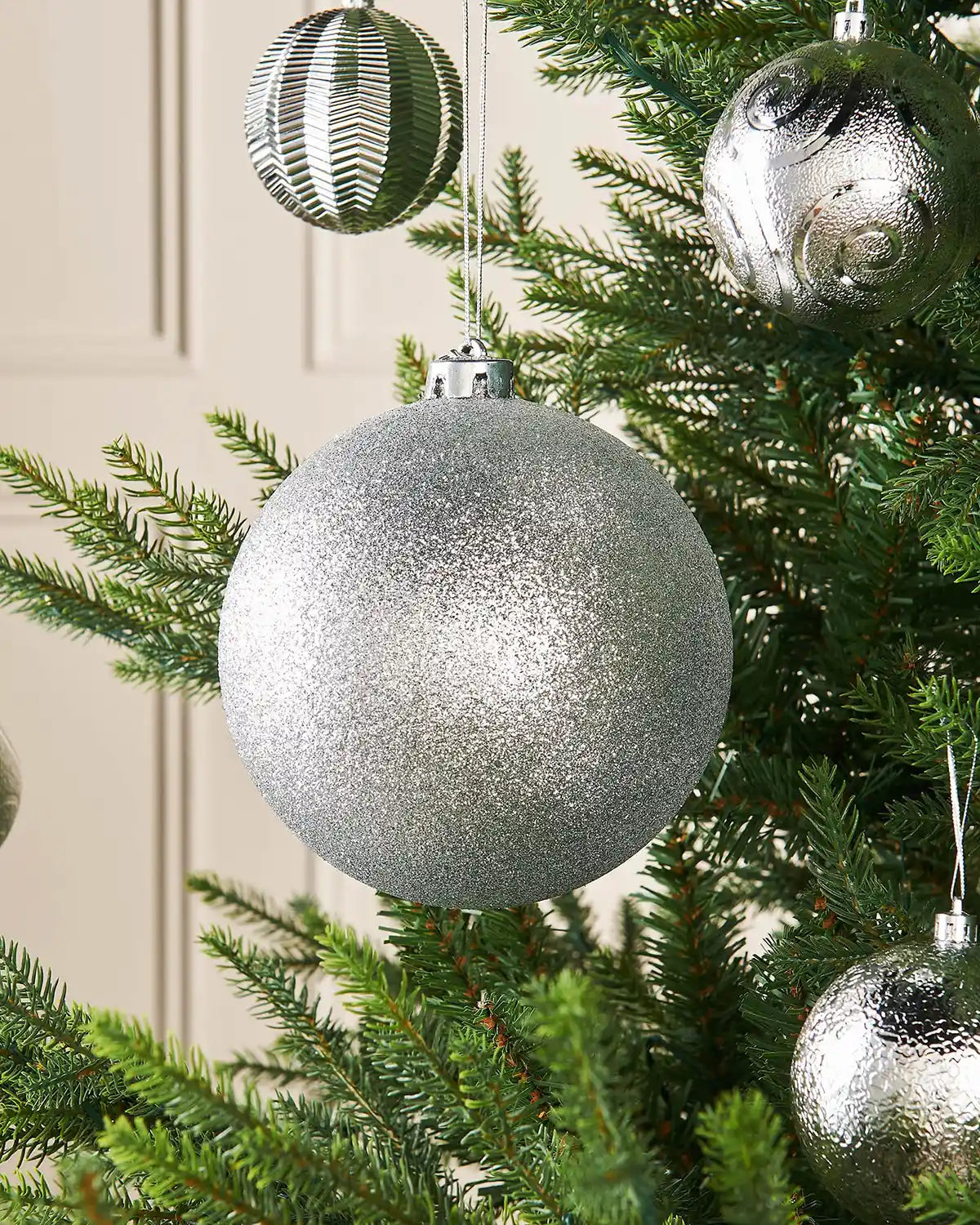 Silver oversized 15cm glitter bauble on a Christmas tree branch with 4 smaller silver baubles in background